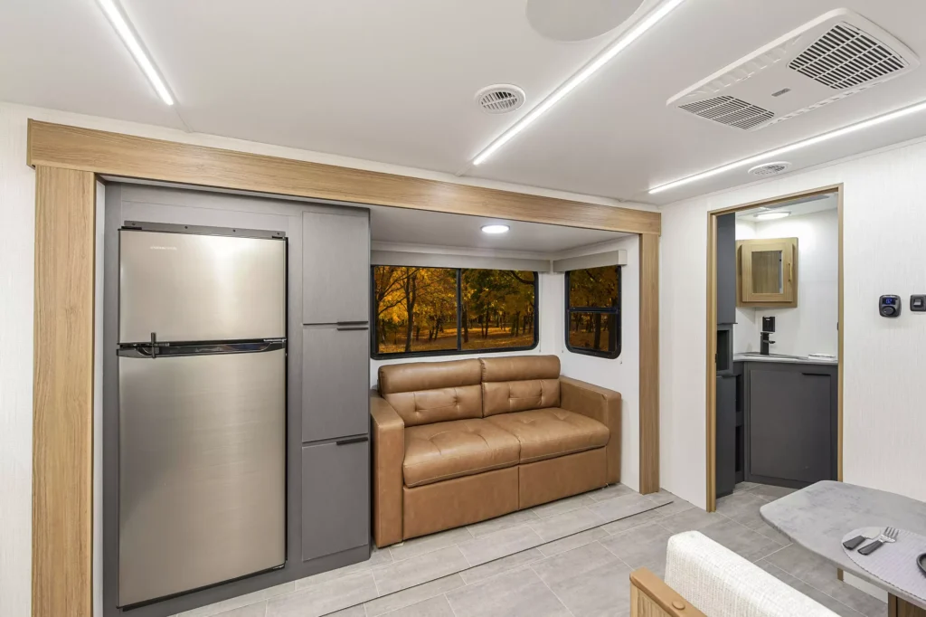 Modern RV interior with a stainless steel refrigerator next to a tan leather sofa. Large windows show a view of autumn trees outside. A compact bathroom and dining area are partially visible.