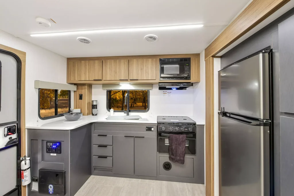 Modern RV kitchen with wood and dark grey cabinets, a refrigerator, oven, microwave, and sink. A window shows autumn trees outside. The space is clean, well-lit, and equipped with various appliances.