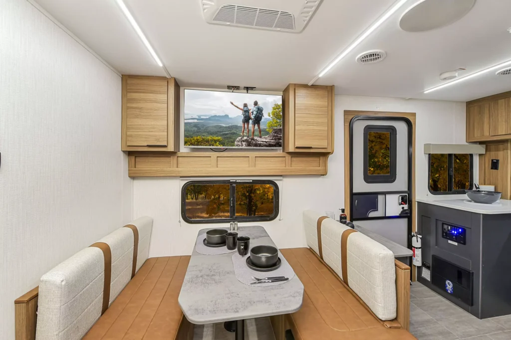 Modern RV interior with a dining area featuring a table set for four, cushioned bench seating, light wood cabinetry, and a kitchen area. A window shows a view of trees and a photo of hikers is displayed above it.