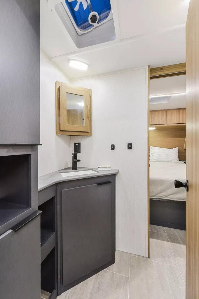 Interior of a modern camper bathroom showing a small sink with a black faucet, mirror cabinet, grey cabinets, and a view into a cozy bedroom with a made bed and wooden headboard.