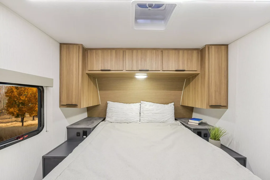 A cozy RV bedroom with a double bed, white and gray bedding, wooden overhead cabinets, nightstands with a book and potted plant, and a window showing an autumn tree outside.