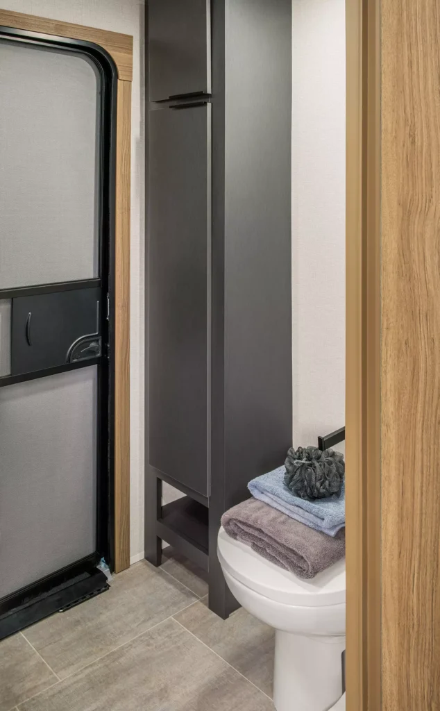 A small, modern bathroom with light wood and gray tones, featuring a white toilet with folded towels on top, a tall dark cabinet, and a black-framed frosted door.