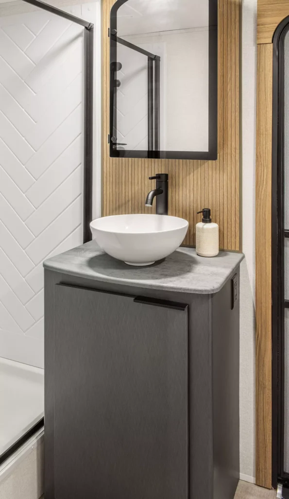 Modern bathroom vanity with a round white vessel sink, black faucet, soap dispenser, and a rectangular mirror above. Wood paneling and a white shower with a chevron tile pattern are partially visible.