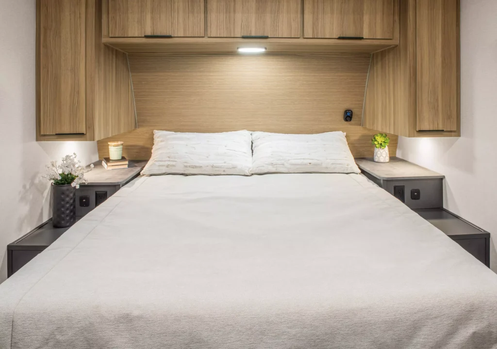 A neatly made bed with white bedding, two pillows, and built-in wooden cabinets above. Nightstands on each side hold small potted plants, a candle, and a cup. A wall light is illuminated above the headboard.