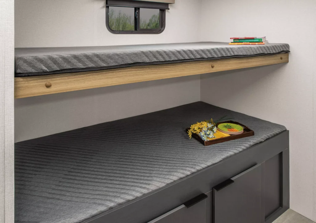 A minimalist room with two gray bunk beds; the top bed holds a stack of books, while the bottom bed has a tray with a meal and flowers. The lower bed has storage drawers underneath. A small window is above the top bed.