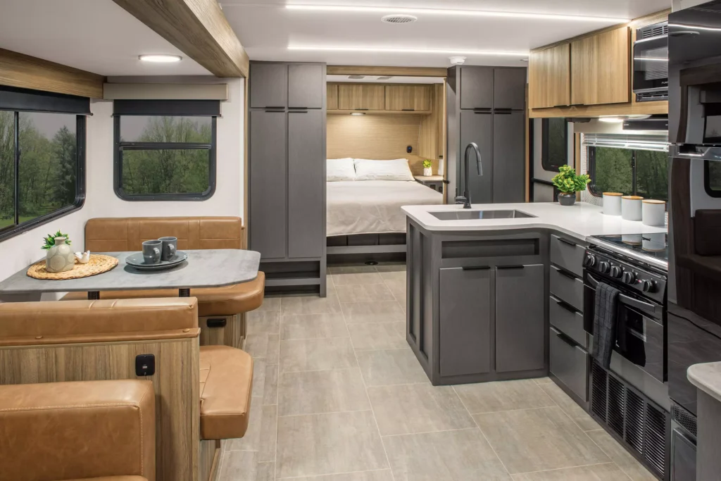Modern RV interior featuring a kitchen with gray cabinets, stove, and sink, a cozy dining area with brown seats, and a bed in the background. Large windows offer views of greenery outside.