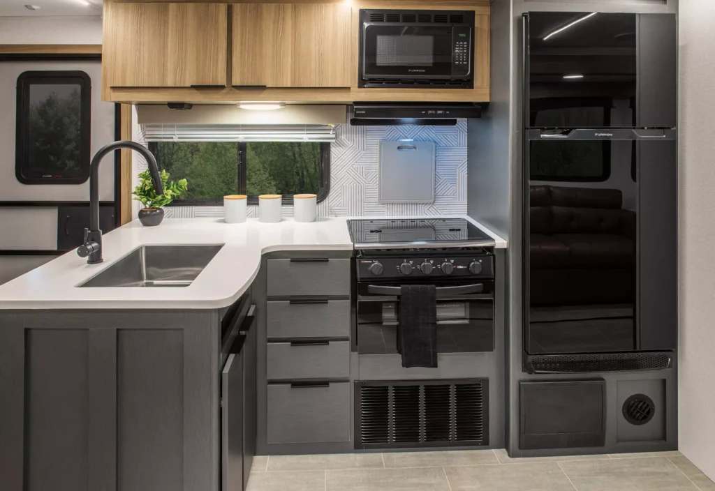 Modern RV kitchen interior featuring a sink with a gooseneck faucet, a stove with oven, microwave, refrigerator, and upper and lower cabinets. A window and plant are above the counter, with neutral decor.