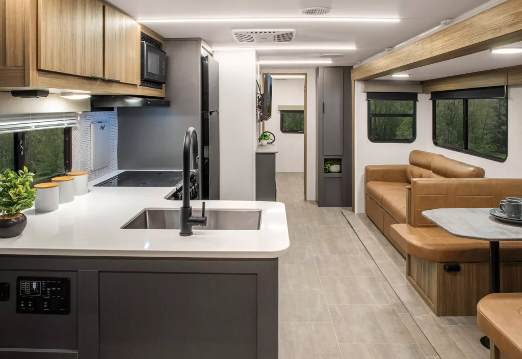 Modern RV interior with a kitchen featuring a sink and stove, wood cabinets, leather sofa, built-in dining area, and large windows showing a green forested landscape outside. The space appears bright, spacious, and well-designed.