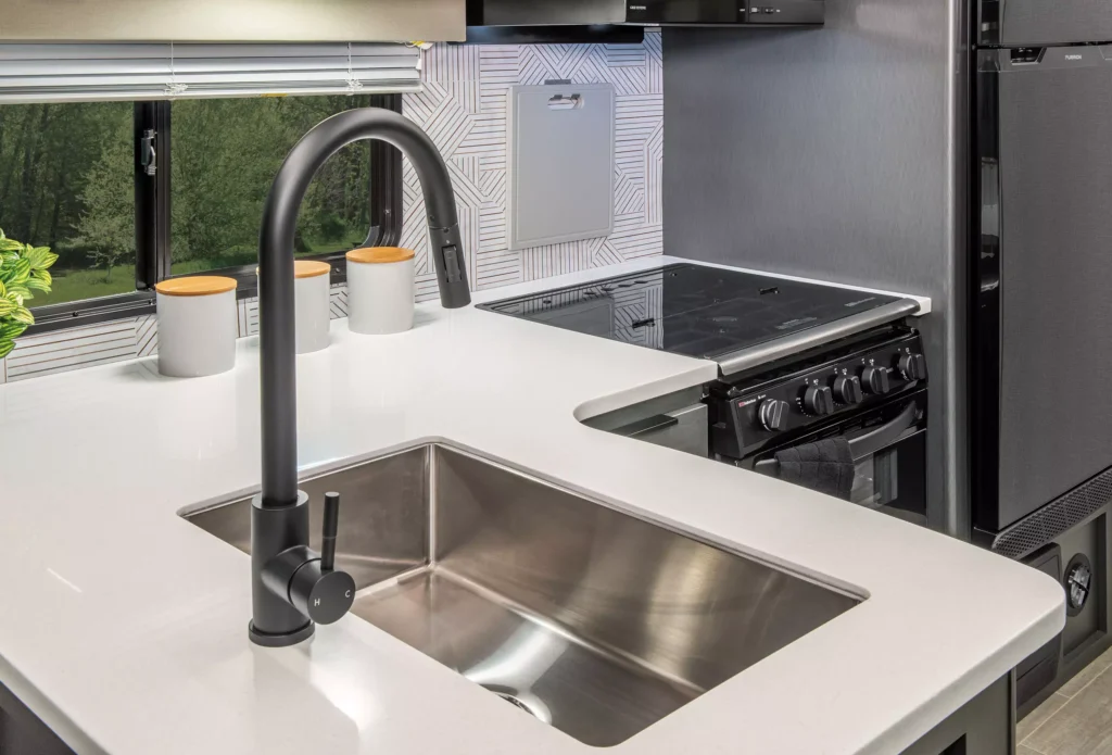Modern kitchen countertop with a stainless steel sink and matte black faucet, near a stove with black knobs, two white canisters, and a large window with a view of greenery.