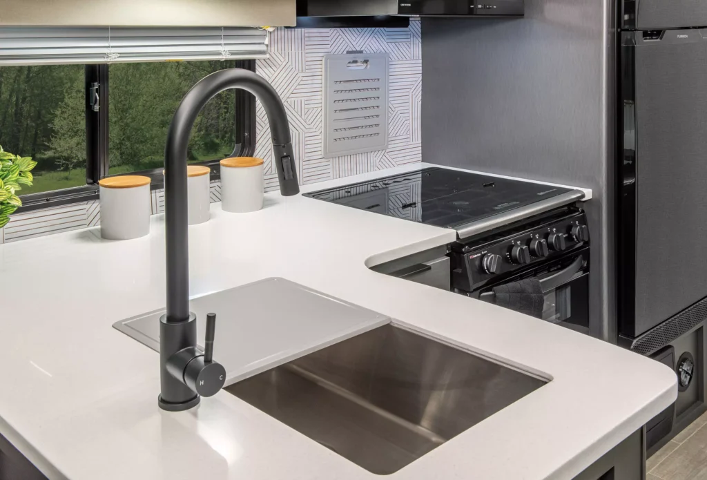 Modern kitchen with a white countertop, stainless steel sink, black faucet, stove, oven, and two white canisters near a window. Geometric backsplash and green scenery visible outside.