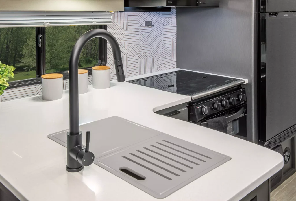 Modern kitchen with a sleek black faucet, a gray sink cover with grooves, a white countertop, two white jars with wooden lids, a patterned backsplash, and a black stove next to a stainless steel refrigerator.