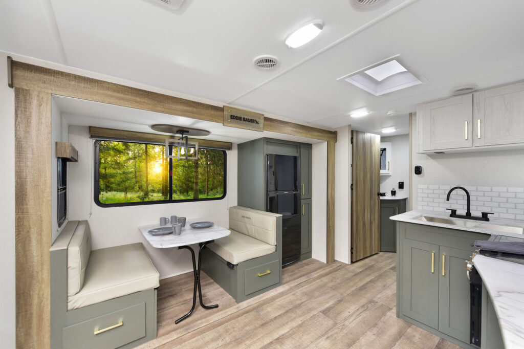 Modern RV interior with light wood floors, green and white cabinetry, a small kitchenette, and a dining area with two benches and a table. Large window shows a forest with sunlight streaming through the trees.
