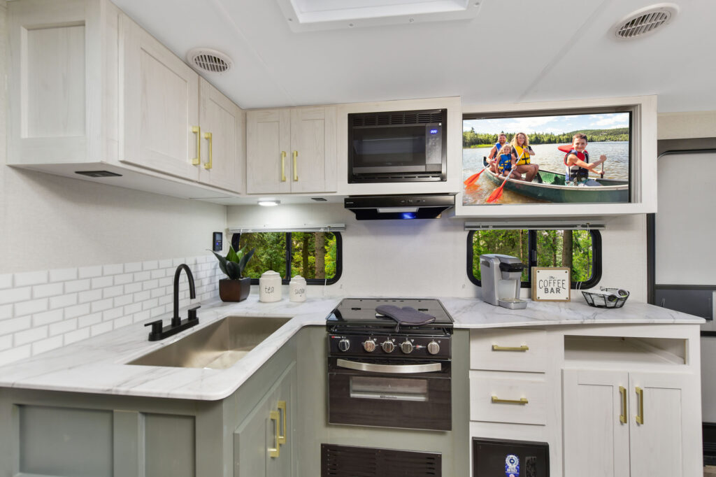 A modern RV kitchen with white cabinets, a black faucet, a stove, microwave, and a coffee bar. A TV above the counter shows two people paddling a canoe on a lake. Greenery is visible through the windows.