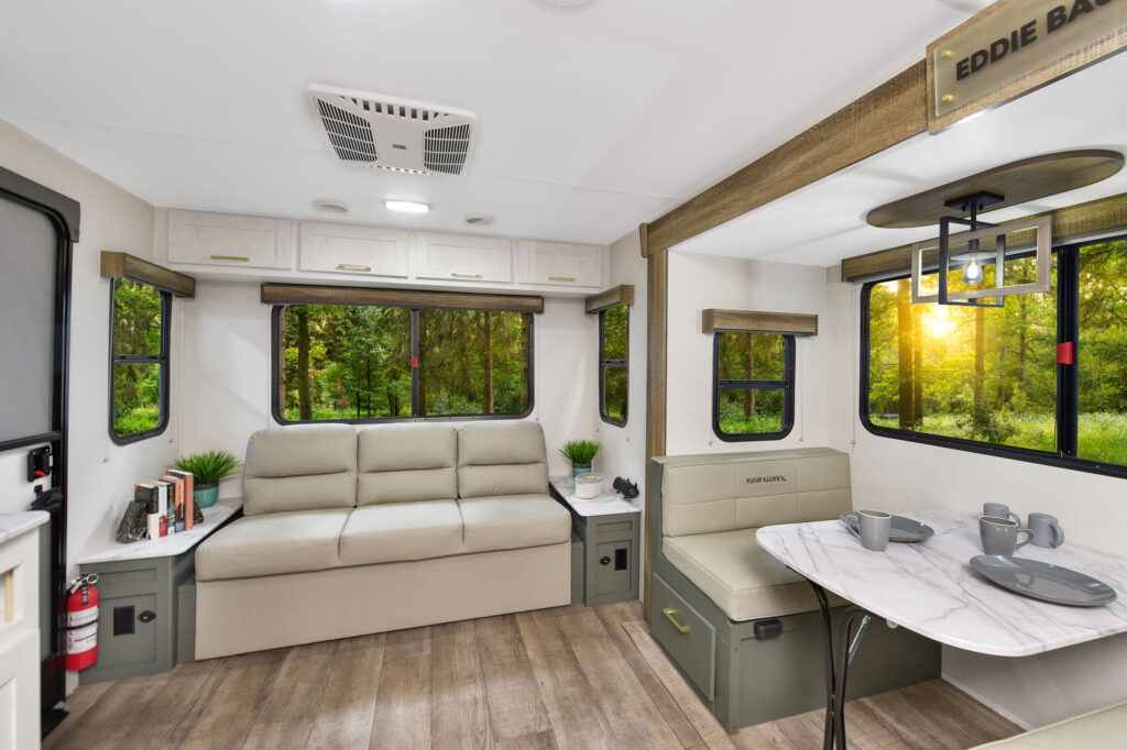 Modern RV interior with beige sofa, dining table set with dishes, large windows showing green trees and sunlight, light wood flooring, built-in storage, and neutral decor. Cozy and inviting atmosphere.