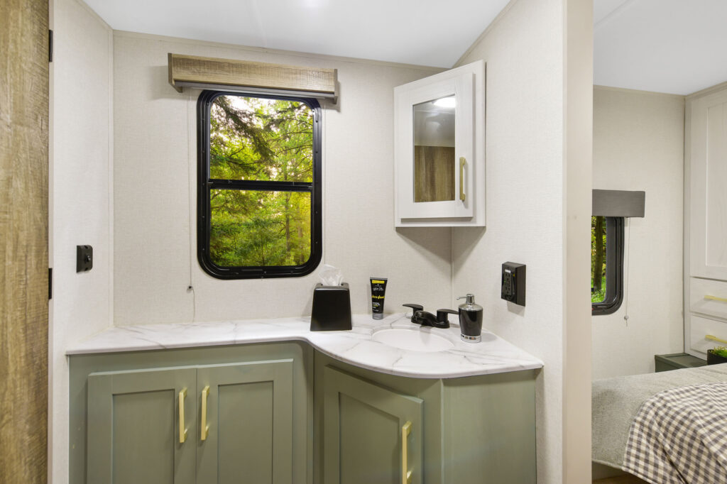 A small bathroom with green cabinets, a white marble counter, a window with a brown shade, a wall cabinet, faucet, soap dispenser, tissues, and grooming products, with trees visible through the window.