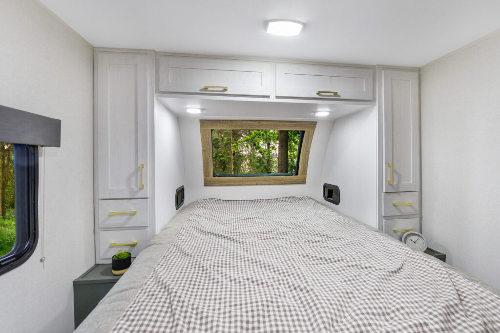 A cozy RV bedroom with a neatly made bed, patterned comforter, overhead cabinets, side windows, and a large rear window showing greenery outside. A small clock and plant sit on bedside shelves.