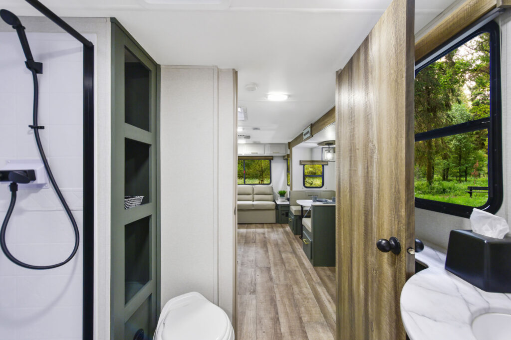 Interior of a modern RV with wood floors, a shower on the left, toilet, vanity with sink, kitchen area, and a sofa at the back; large windows show green trees and natural light streams inside.