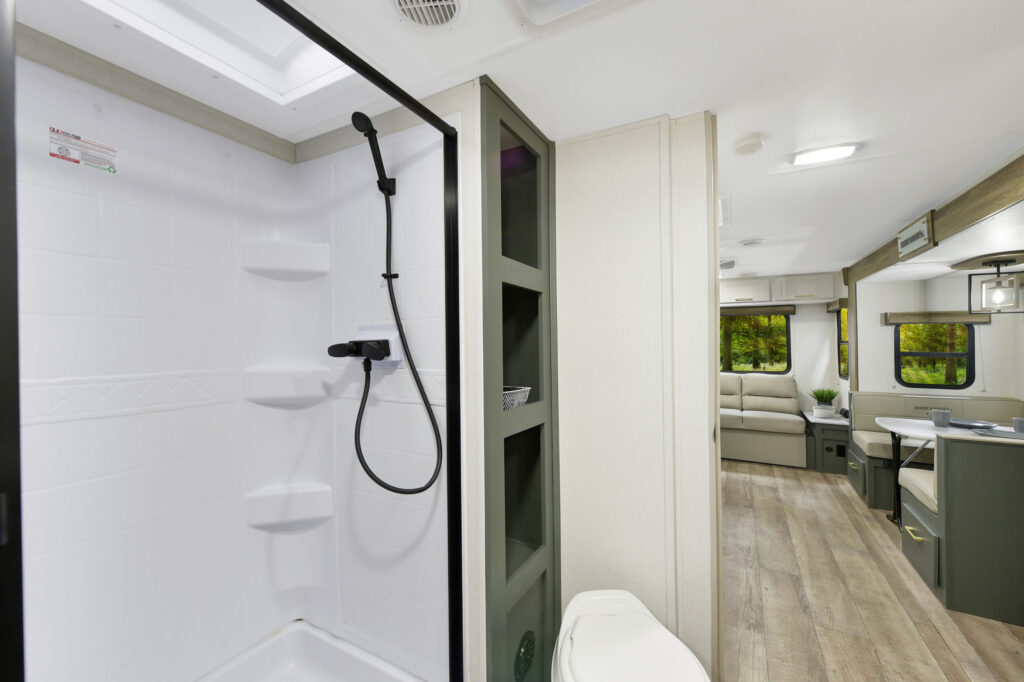 Interior of a modern RV showing a white shower with built-in shelves and a black showerhead, a toilet, and a view into a living area with seating, large windows, and laminate wood flooring.