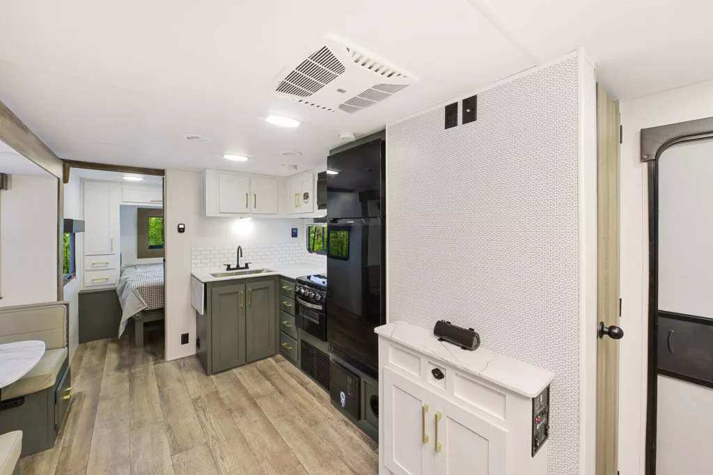 Modern RV interior with light wood floors, a small kitchen with green cabinets, a dining area, and a bedroom visible in the back. White walls and ceilings create a bright, open atmosphere.