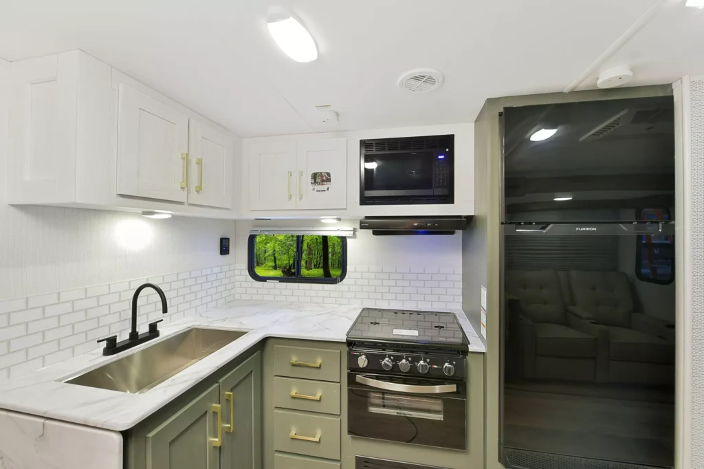 Modern RV kitchen with white upper cabinets, sage green lower cabinets, gold hardware, a double sink, black faucet, white subway tile backsplash, black stove with oven, microwave, and large refrigerator. Window shows greenery outside.