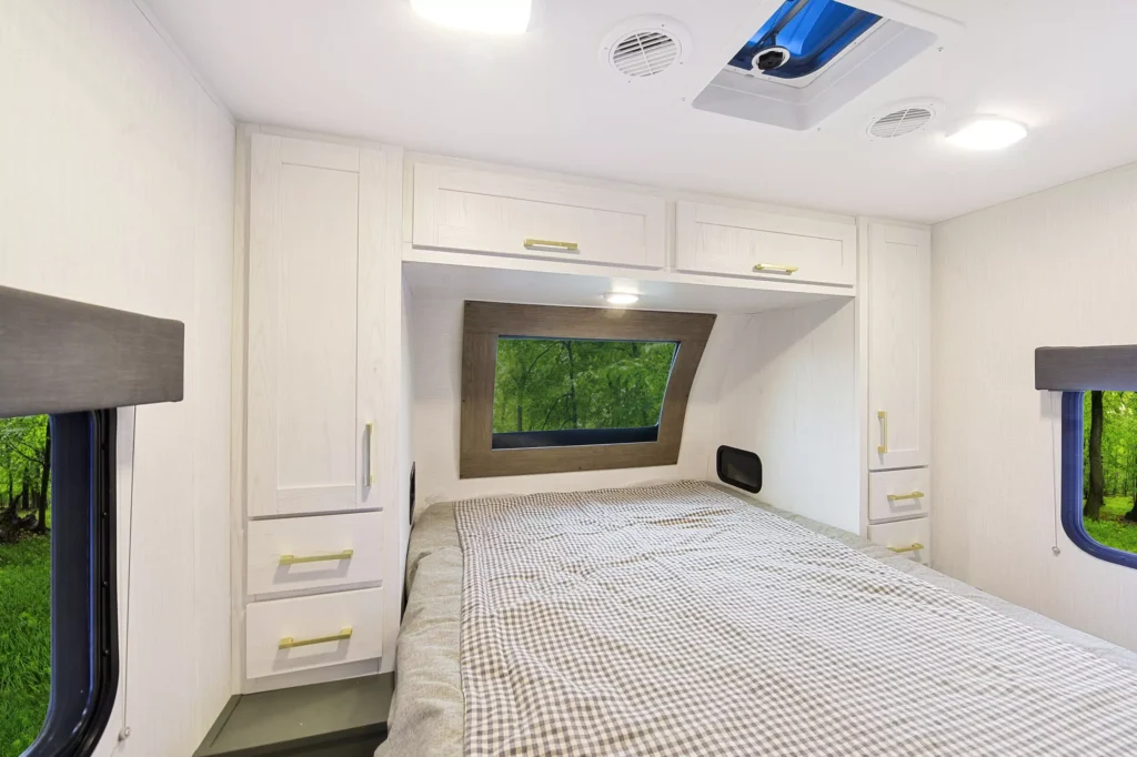 Interior of a modern RV bedroom with built-in white cabinets, a large window above the head of a neatly made bed, and another window on the left, showing green trees outside. Ceiling lights and vents are visible.