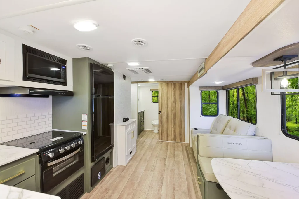 Bright, modern RV interior featuring a kitchen with a stove and microwave, a dining area with a cushioned bench, wood-style flooring, large windows with views of greenery, and a door leading to a bathroom.