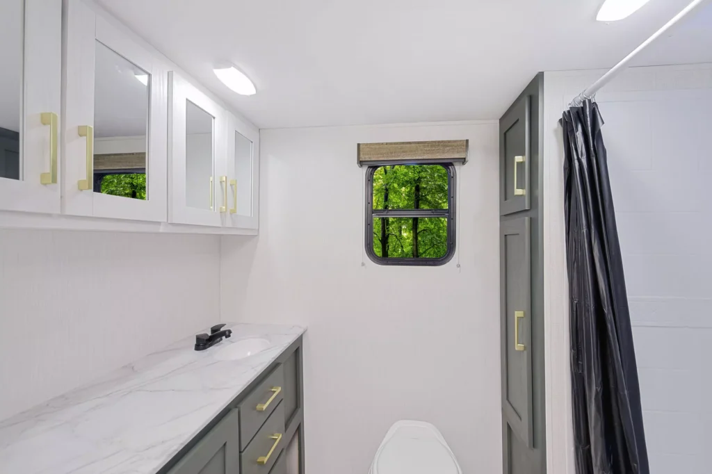 Modern bathroom with marble countertop, gray and white cabinets, gold handles, a small window showing green trees outside, a toilet, and a shower with a dark curtain on the right.