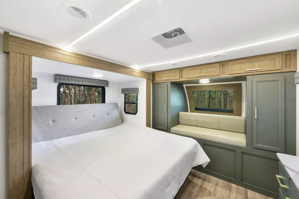 Modern RV bedroom with a large bed, padded headboard, wood and green cabinetry, overhead storage, and a built-in bench by a large window overlooking a forest. Natural light fills the room.