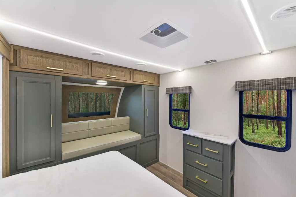Modern RV bedroom interior with wood cabinets, a cushioned bench under a window, green drawers, two windows displaying a forest view, and a white ceiling with recessed lighting.