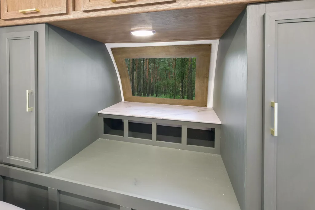 A built-in gray desk nook with upper and side cabinets, a white surface, and a large window offering a view of a dense green forest. A round ceiling light is mounted above the desk area.