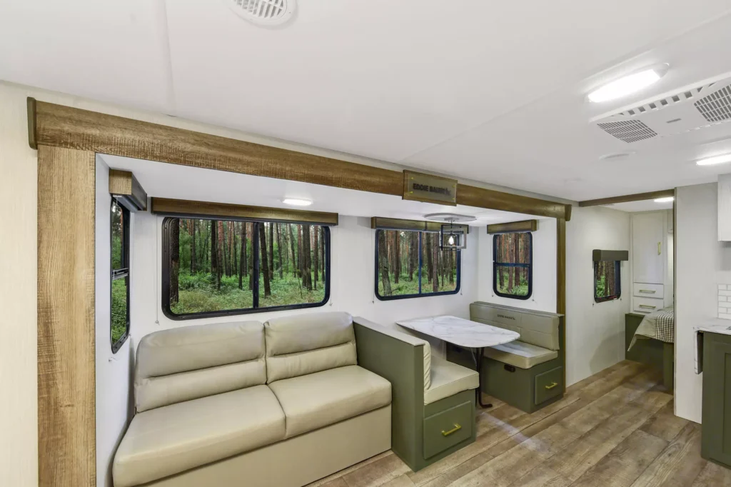 Interior of an RV featuring a beige sofa, a dining area with a table and benches, large windows showing a forest outside, and wood-look flooring. The space appears clean and modern with neutral colors.