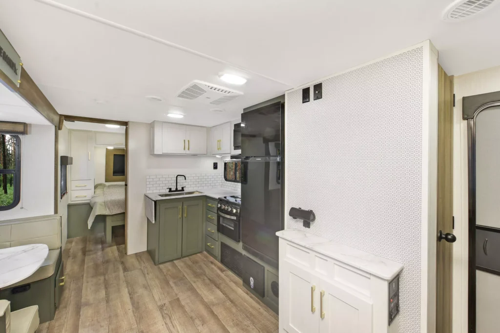 Modern RV interior with wood-look flooring, a compact kitchen featuring green lower cabinets, white upper cabinets, and black appliances. A small dining area and a bed are visible in the background. Light, airy design.