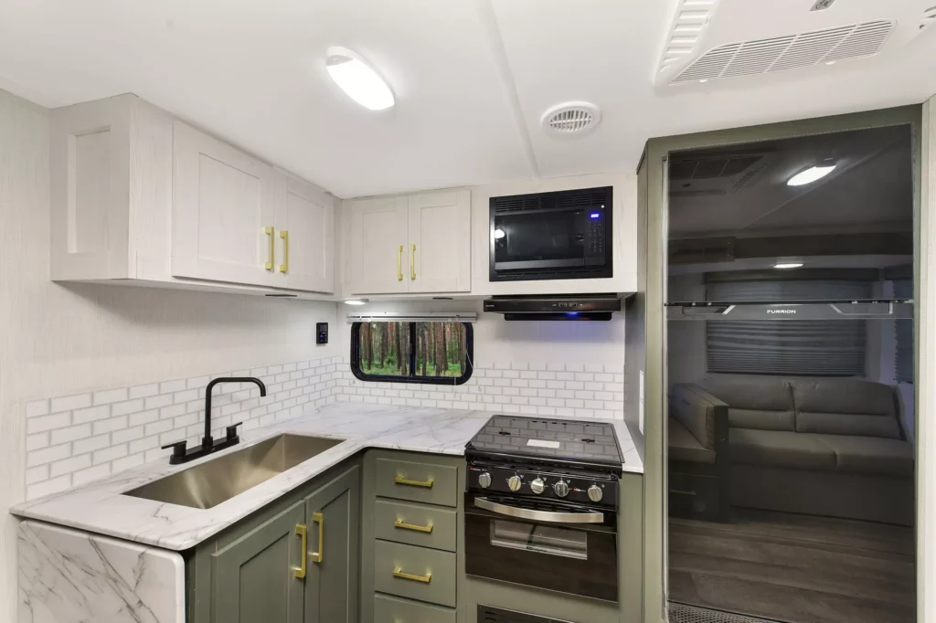 Modern RV kitchen with marble countertops, farmhouse sink, green and white cabinetry, a black faucet, gas stove, built-in microwave, white tile backsplash, and a view of a sofa in an adjoining room.