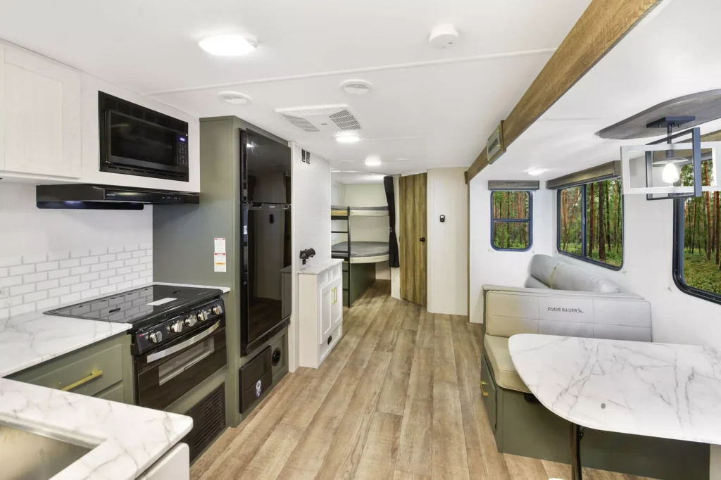 Interior of a modern RV showing a kitchen with stove and microwave, a dining area with cushioned benches and a table, wood-style flooring, large windows, and a sleeping area with bunk beds in the back.