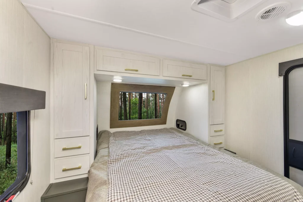 Interior of an RV bedroom with white cabinets above and beside a bed, plaid bedding, windows on two sides showing a forest view, and neutral-colored walls and ceiling.