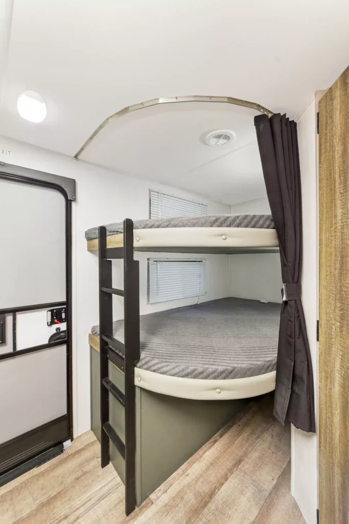 Two bunk beds with gray mattresses and black frames are built into a small, clean RV interior. A black curtain on the right can be pulled for privacy. A door and window are visible on the left, with wood-style flooring below.