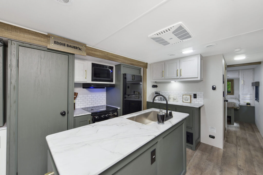 Modern RV kitchen with green cabinets, white marble countertops, a center island with a sink, oven, microwave, and white tile backsplash; a hallway leads to a bedroom in the background.
