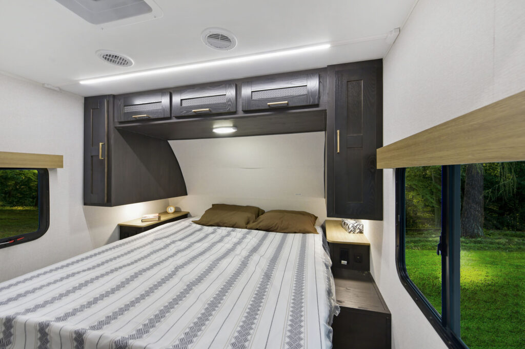Interior of an RV bedroom featuring a bed with striped bedding and brown pillows. Dark wood overhead cabinets provide storage. A window to the right offers a view of green foliage outside. Neutral walls and ceiling lights enhance the cozy space.