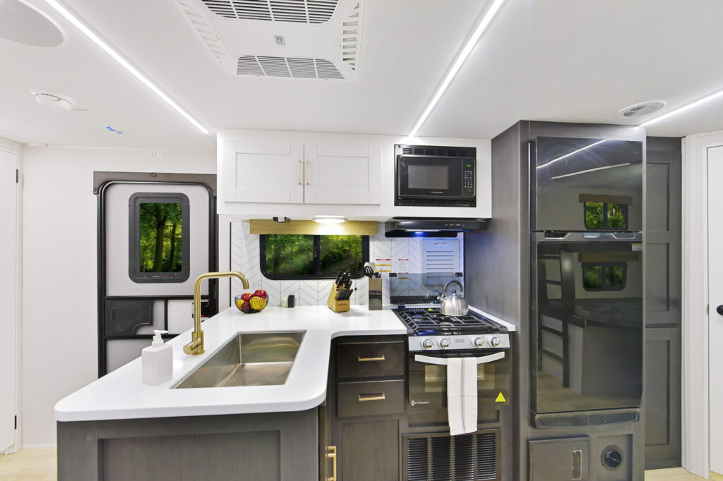 Modern RV kitchen with sleek white counters, a stainless steel sink, stove with kettle, microwave, and fridge. A fruit bowl sits on the counter, and a window reveals a view of green trees. LED strip lights illuminate the space.