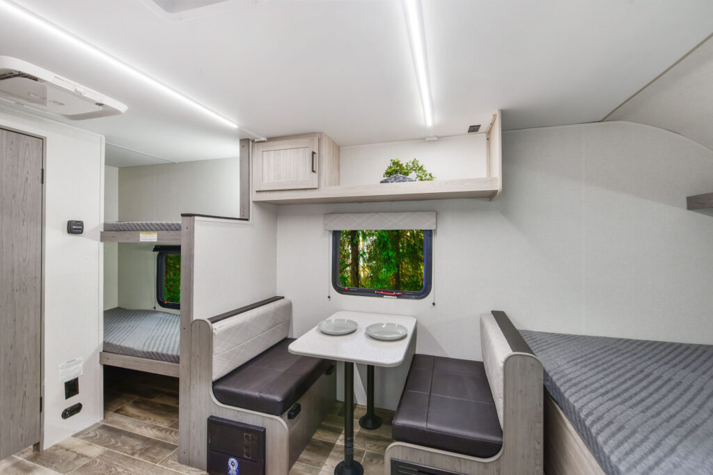 Modern RV interior with a small dining area featuring a table and two benches, bunk beds to the left, and a window showing greenery outside. Light wood and gray tones create a bright, clean look.