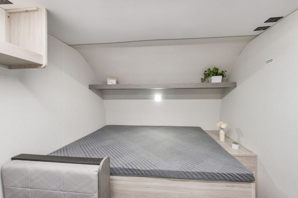 A minimalist bedroom with a neatly made bed, light wood furniture, gray bedding, a floating shelf holding a clock and plant, and soft lighting against light-colored walls.