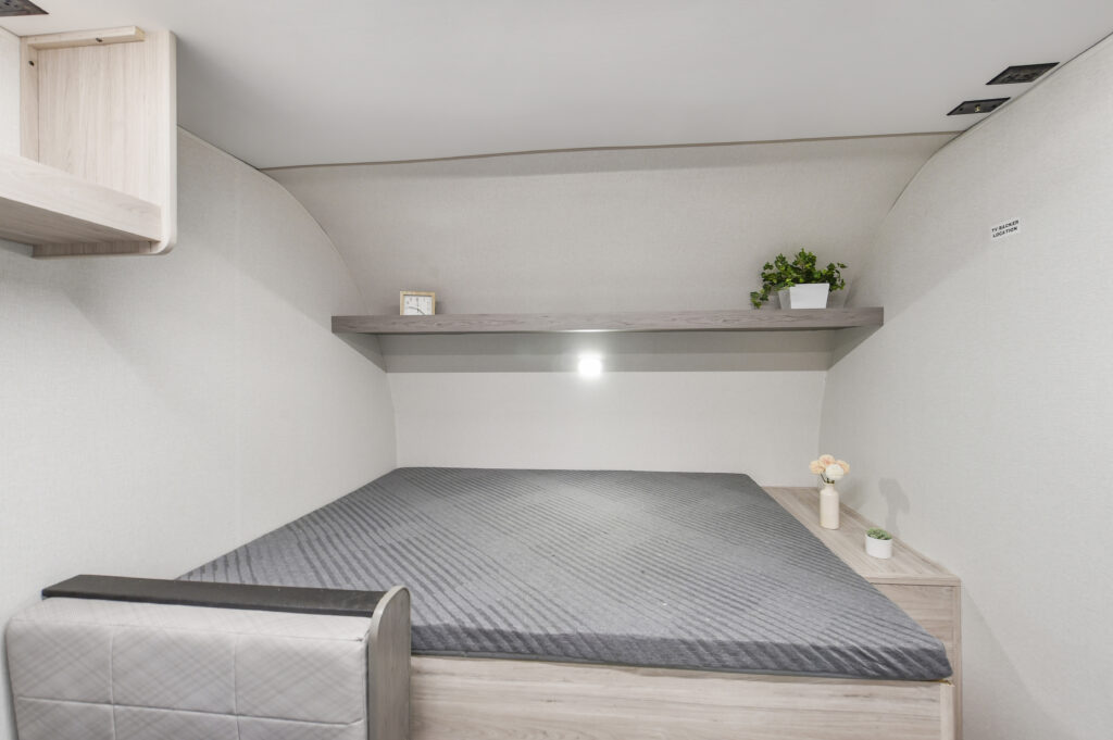 Minimalist bedroom with a gray mattress, built-in light, shelf above the bed holding a potted plant and decor, light wood furniture, and neutral walls.