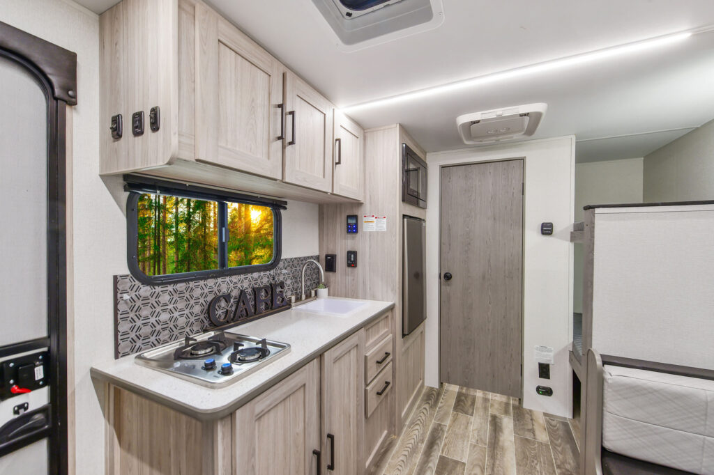 Modern RV kitchen interior with light wood cabinets, a sink, a two-burner stove, backsplash, and a window showing a forest view at sunset. Door and additional storage visible in the background.
