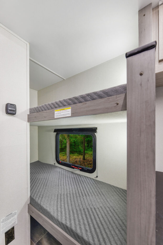 A small bunk bed area inside an RV, featuring two twin mattresses with gray striped covers, a window showing trees outside, and light-colored walls with a black switch on the left.