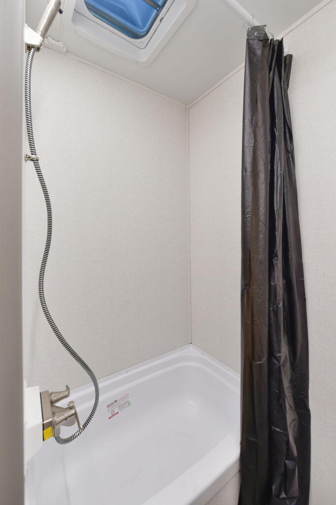 Small shower area with a white tub, simple wall paneling, a black shower curtain, a flexible showerhead, and a ceiling vent with a blue-tinted cover letting in light.