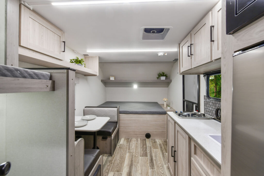 Interior of a modern RV with light wood cabinets, a small dining area, a bed in the back, compact kitchen with stove, sink, and refrigerator, and a window showing greenery outside.