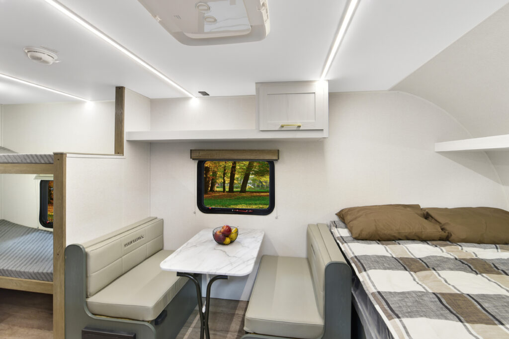 Modern RV interior with a small dining table and two benches, a bed with plaid blankets and pillows, bunk beds, and a window showing a green park scene with trees and sunlight. A bowl of fruit sits on the table.