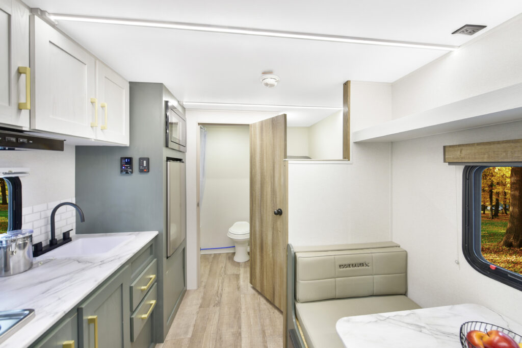 Modern RV interior with a compact kitchen, sink, stove, refrigerator, and dining area. Light wood flooring and neutral cabinetry create a bright atmosphere. An open door reveals a small bathroom with a toilet at the rear.