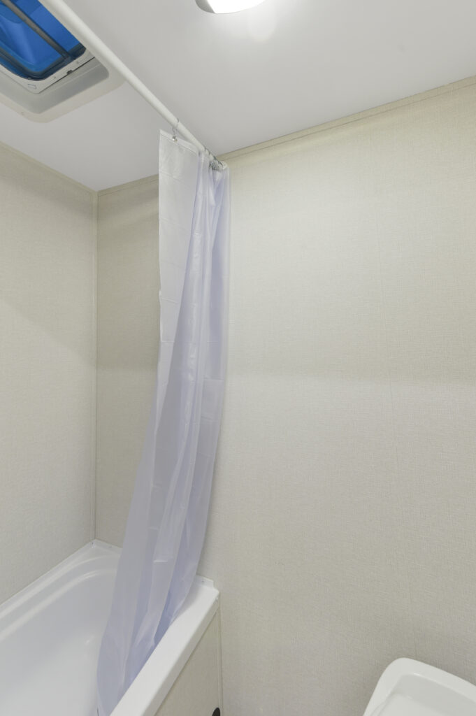 A simple bathroom with a white bathtub, a plain white shower curtain hanging from a rod, light-colored walls, and a ceiling vent with a blue cover.