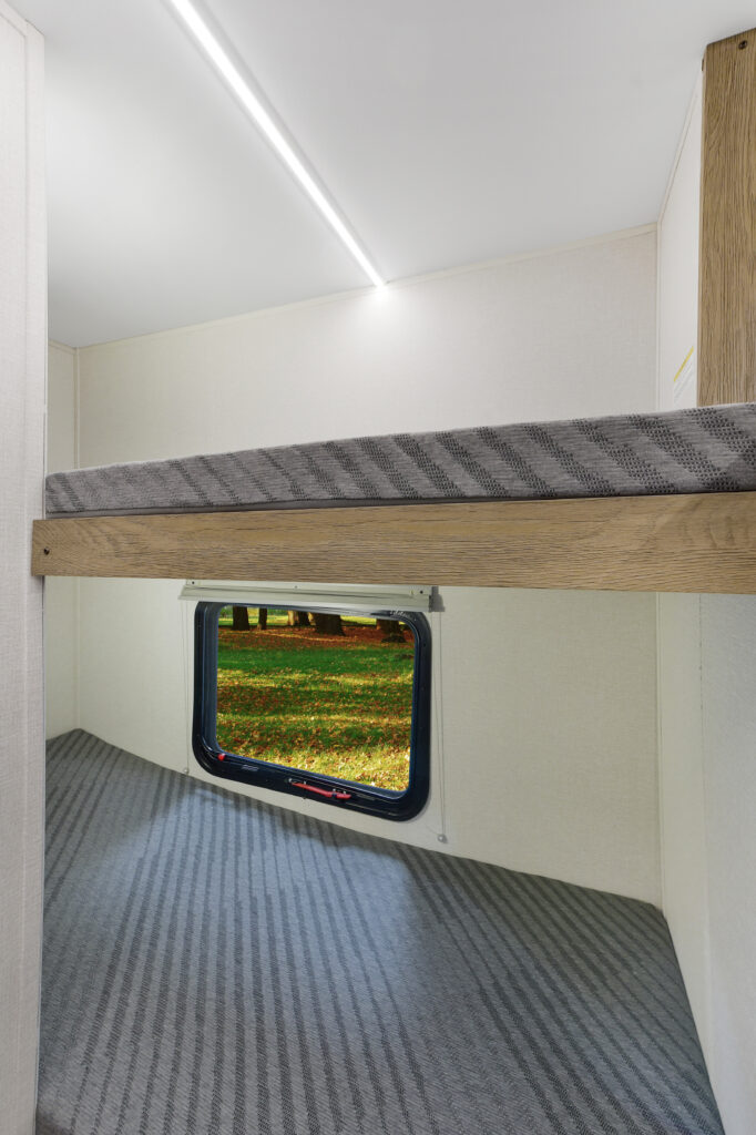 Interior of a camper van showing a small bunk bed with wood accents, striped gray mattresses, and a window overlooking a grassy, sunlit outdoor scene with scattered autumn leaves.